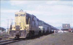 A diesel locomotive with passenger cars stretching behind it waits beside a small red depot building. 